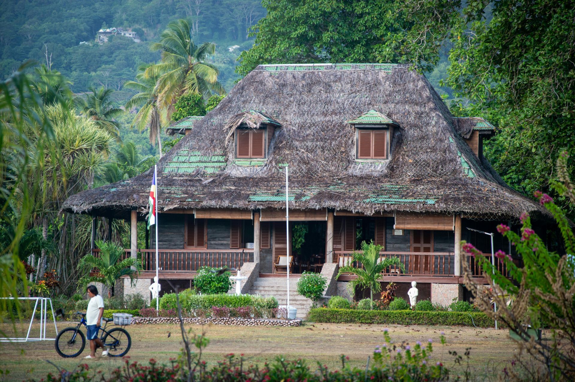 A large, thatched-roof house with a porch, lush greenery, and a person on a bicycle in a yard. Seychelles