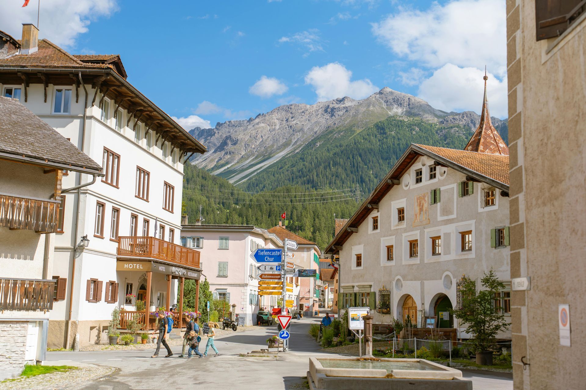 Street view in a European village with buildings, mountains in the background, and pedestrians.
