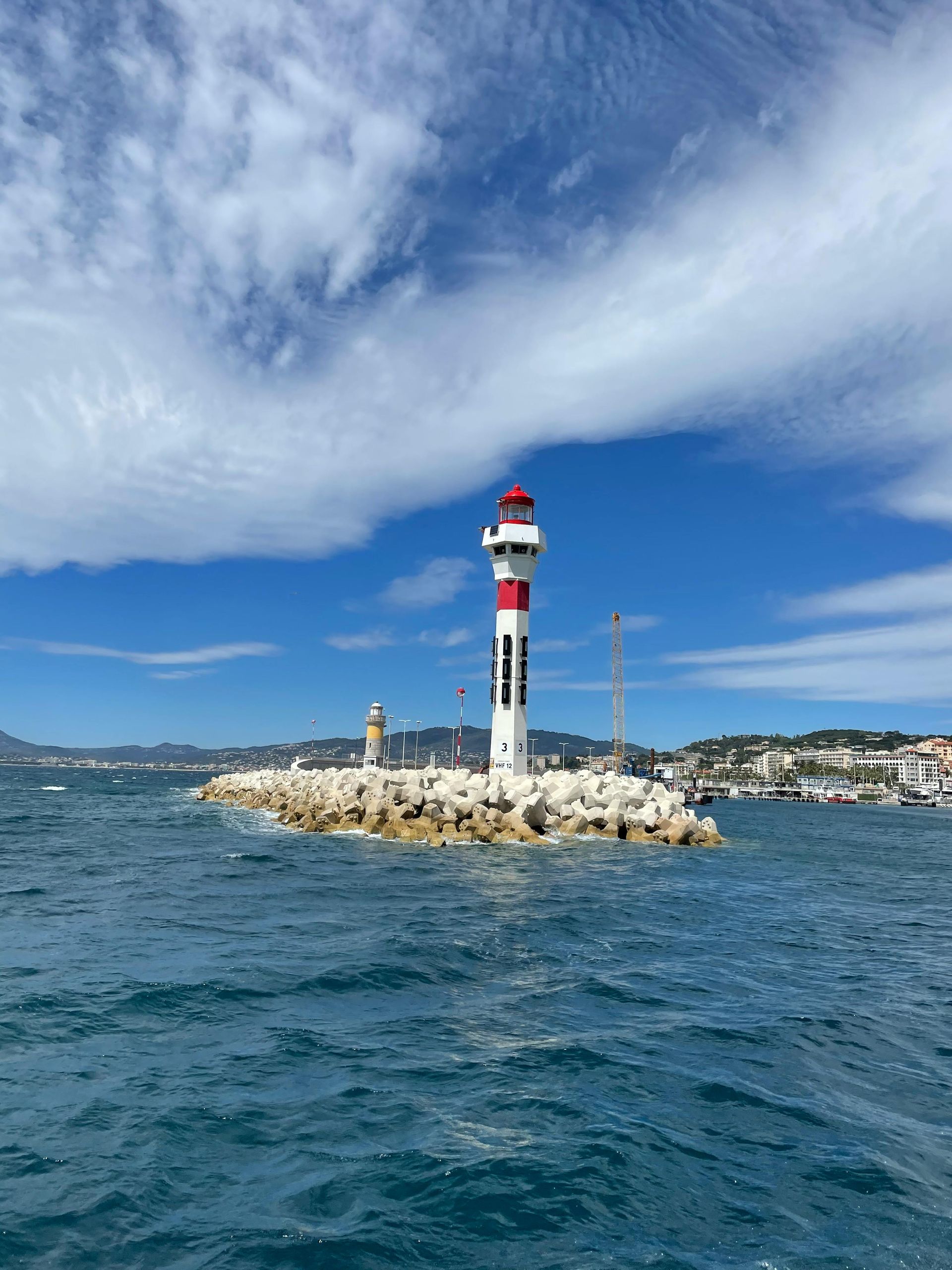 Lighthouse on a rocky breakwater in turquoise water under a blue sky with fluffy clouds. Cap d’Antibes