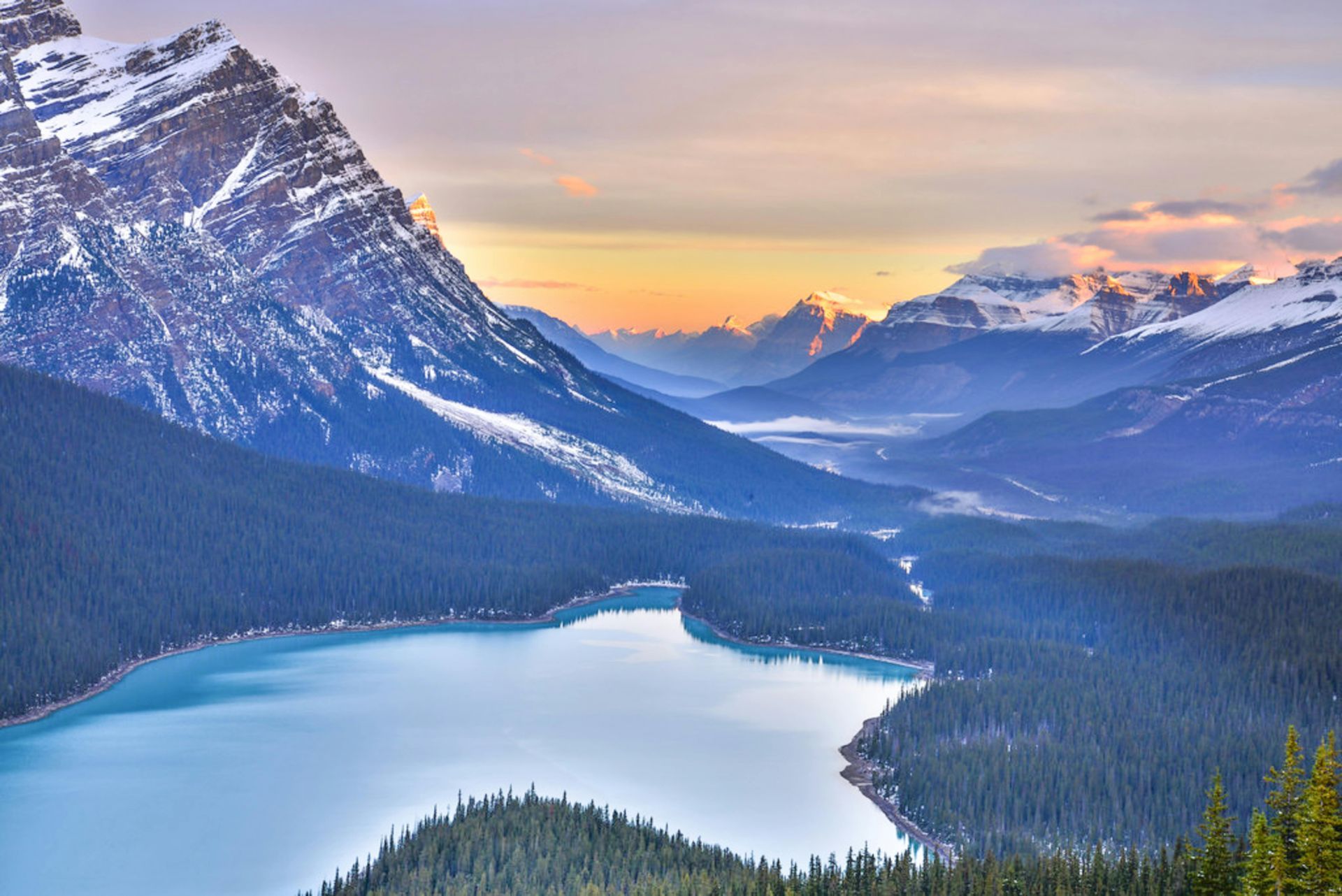 A scenic, high-angle view of a turquoise glacial lake surrounded by snowy mountain peaks under a soft, golden sunrise.