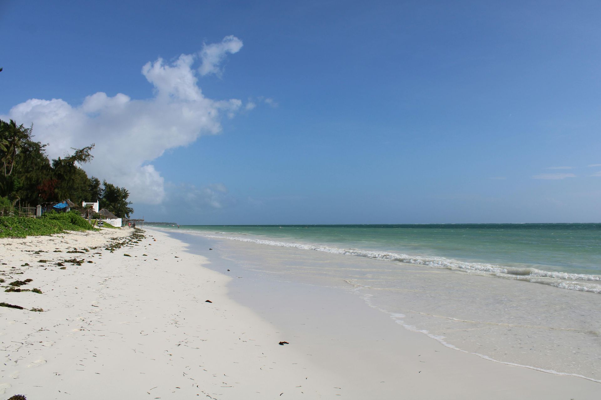 A serene, white sand beach stretches along a calm, turquoise ocean under a bright blue sky with scattered white clouds.