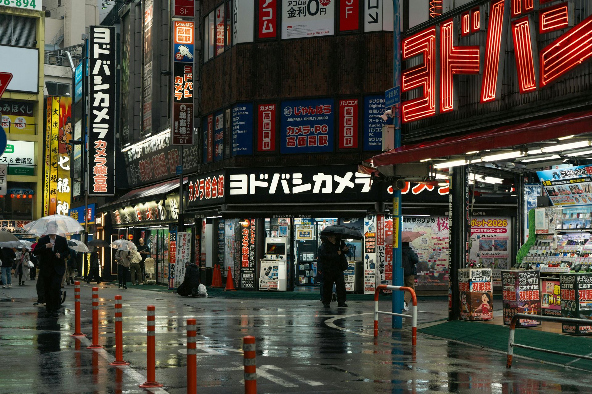 A rainy street scene in a Japanese city, featuring a Yodobashi Camera store with illuminated neon signs at dusk.