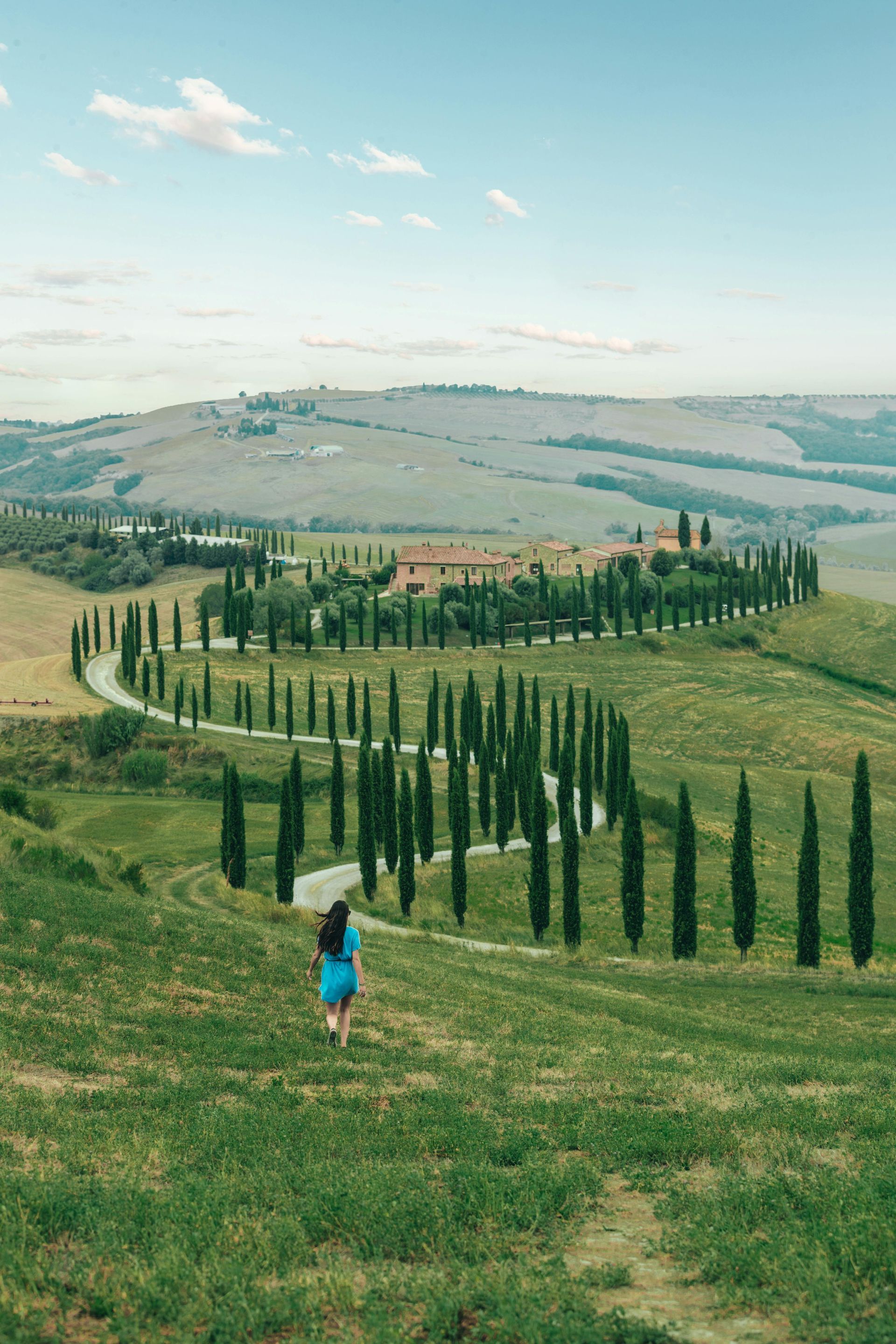Woman walking on a grassy hill towards a winding road lined with cypress trees leading to a building; blue sky.