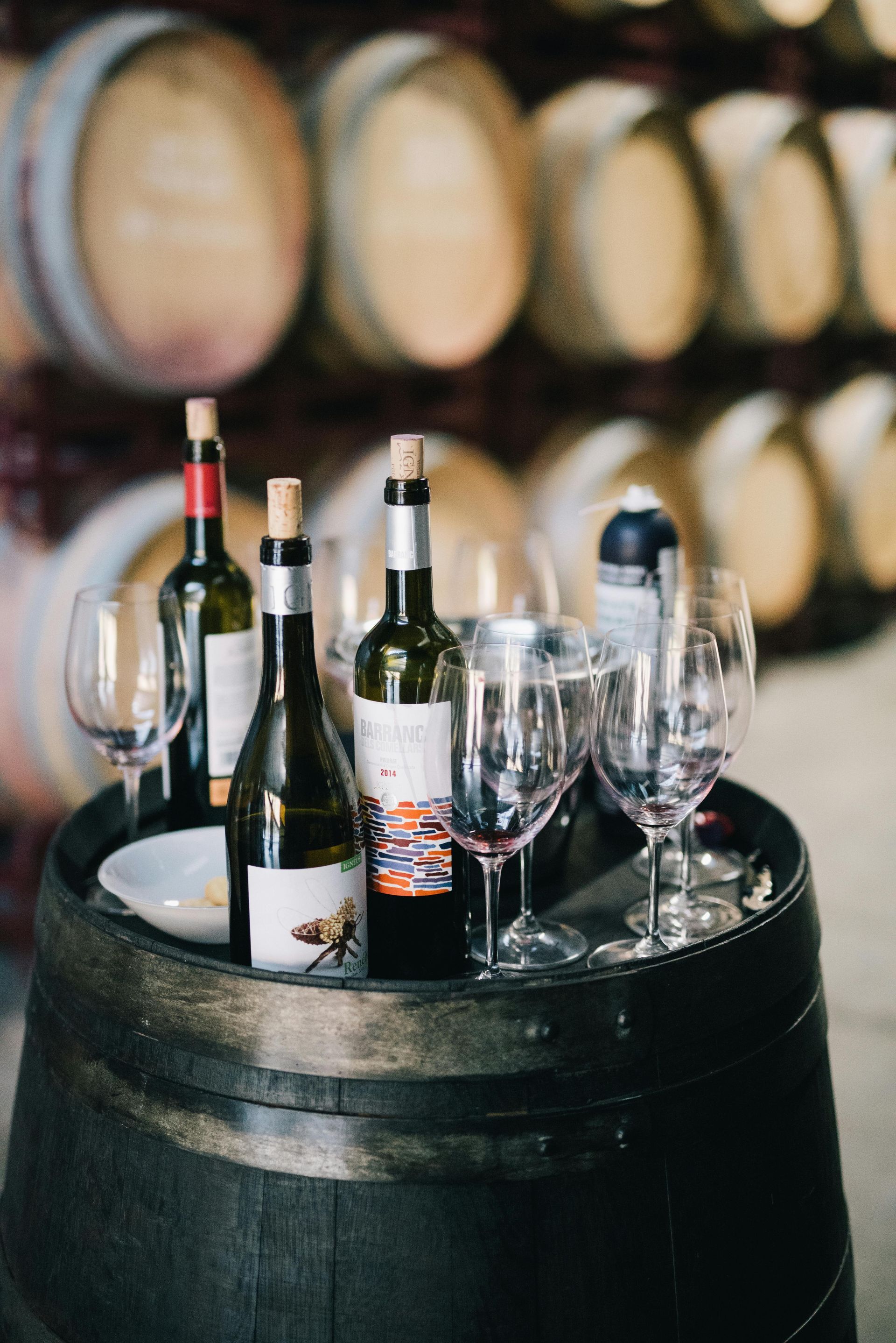 Wine bottles and glasses sit on a wooden barrel in a cellar with stacked wine barrels in the blurred background.