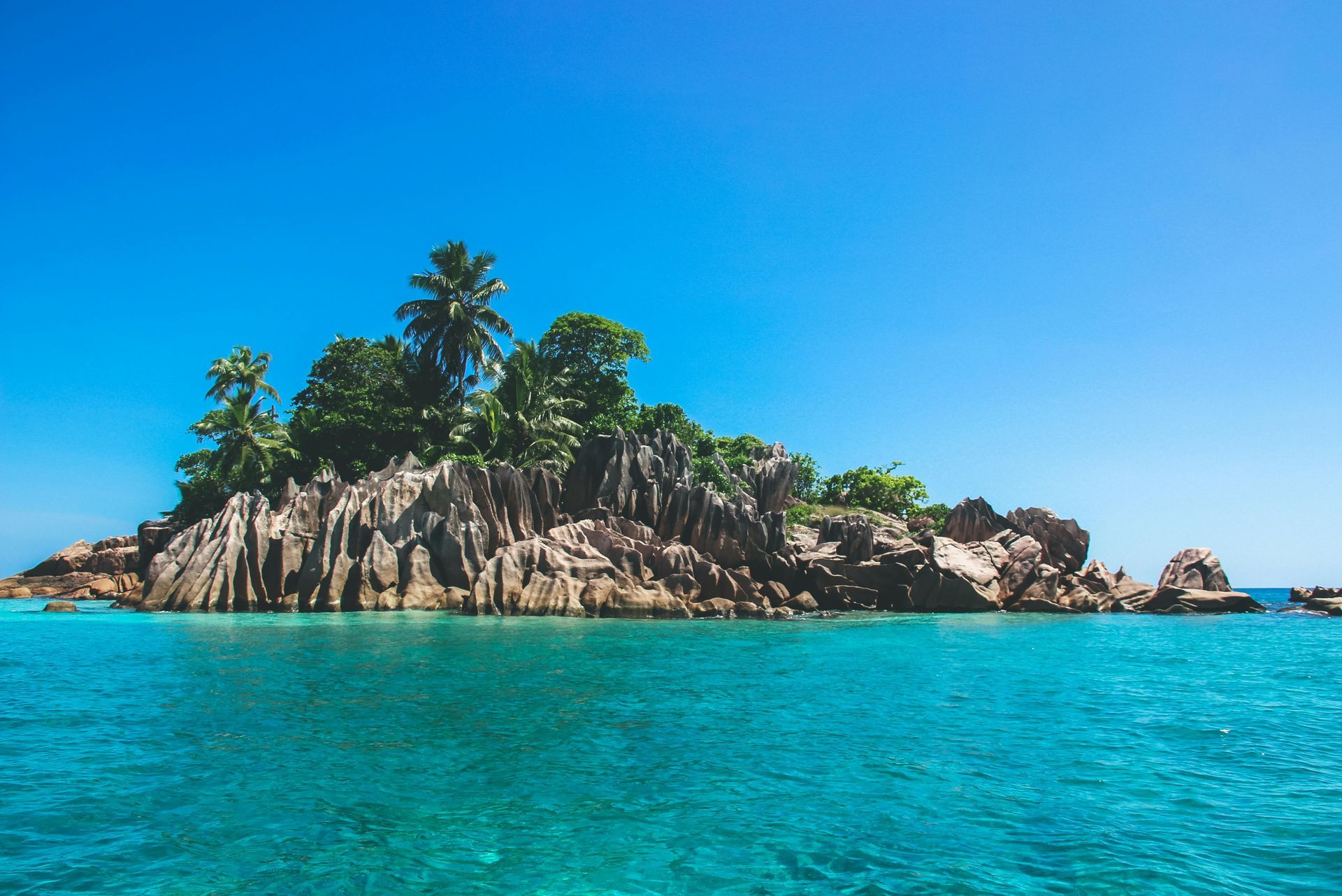 Island with rock formations and palm trees in turquoise water under a clear blue sky. Seychelles