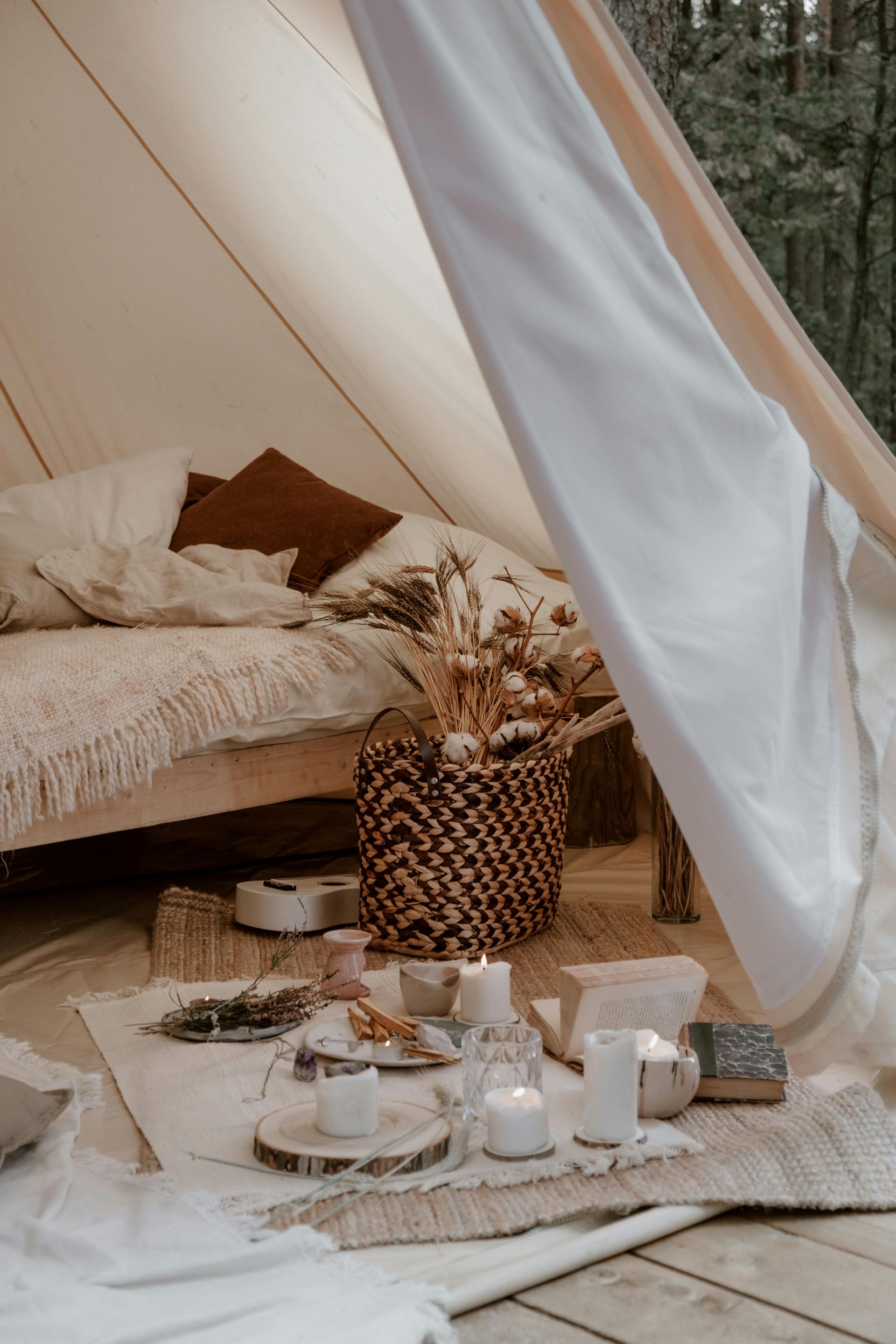 Cozy tent interior with bed, basket of dried flowers, candles, and wooden accents, set in a forest.