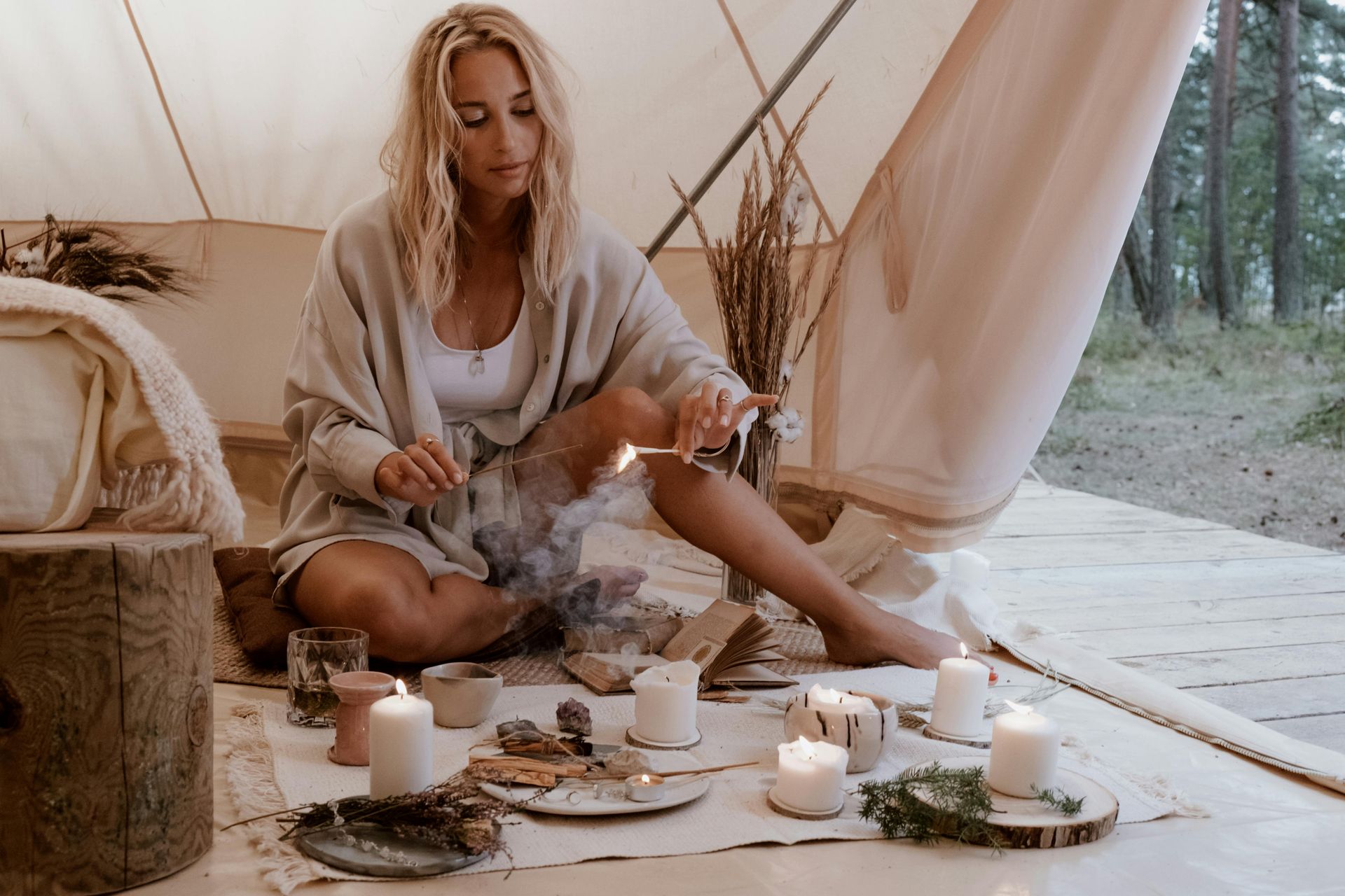 Blonde woman in beige shawl lights incense in tent. Candles and crystals on table, forest backdrop.
