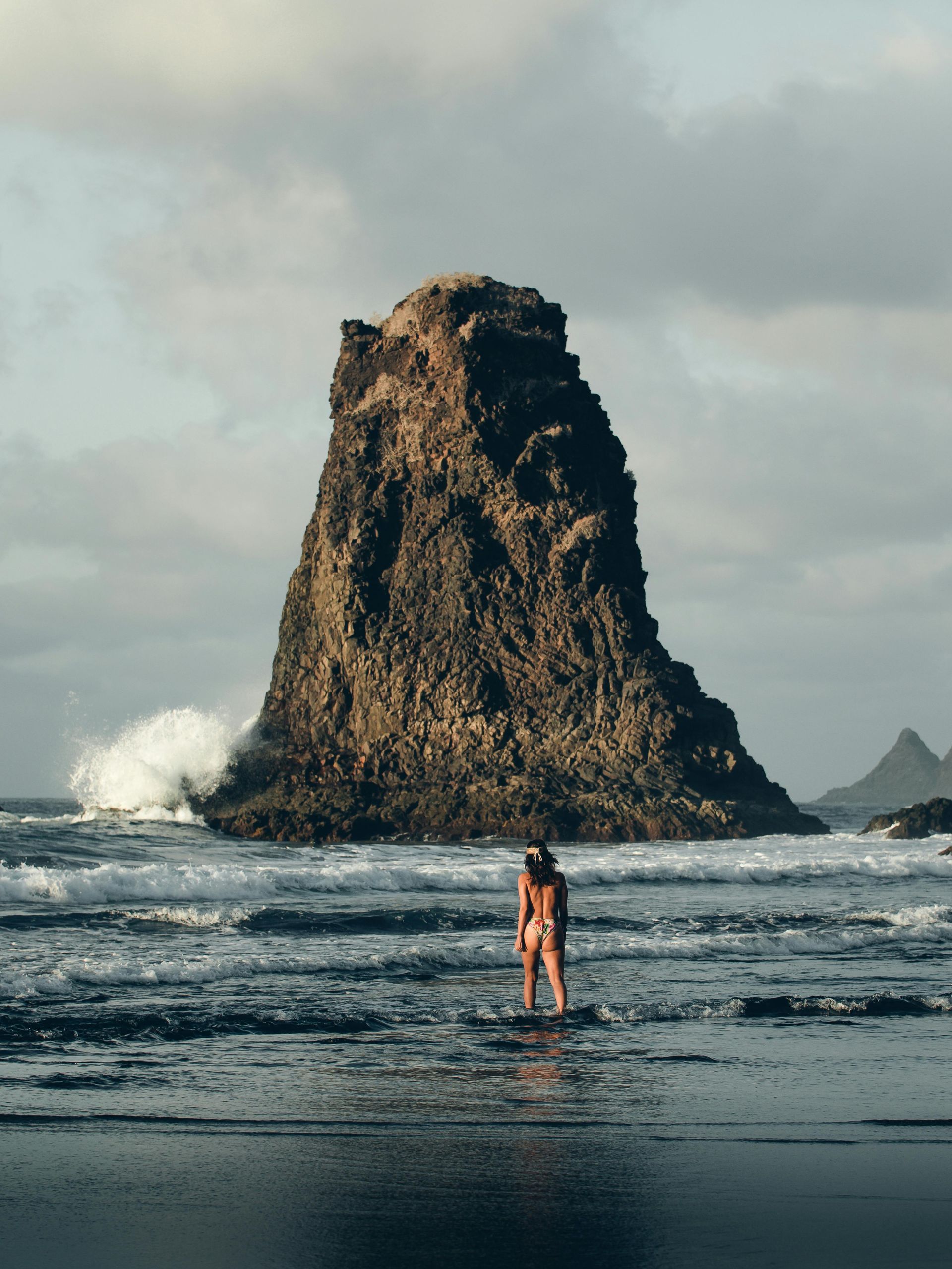 A person stands in shallow ocean water, facing a large, dark, jagged sea stack under a cloudy sky.