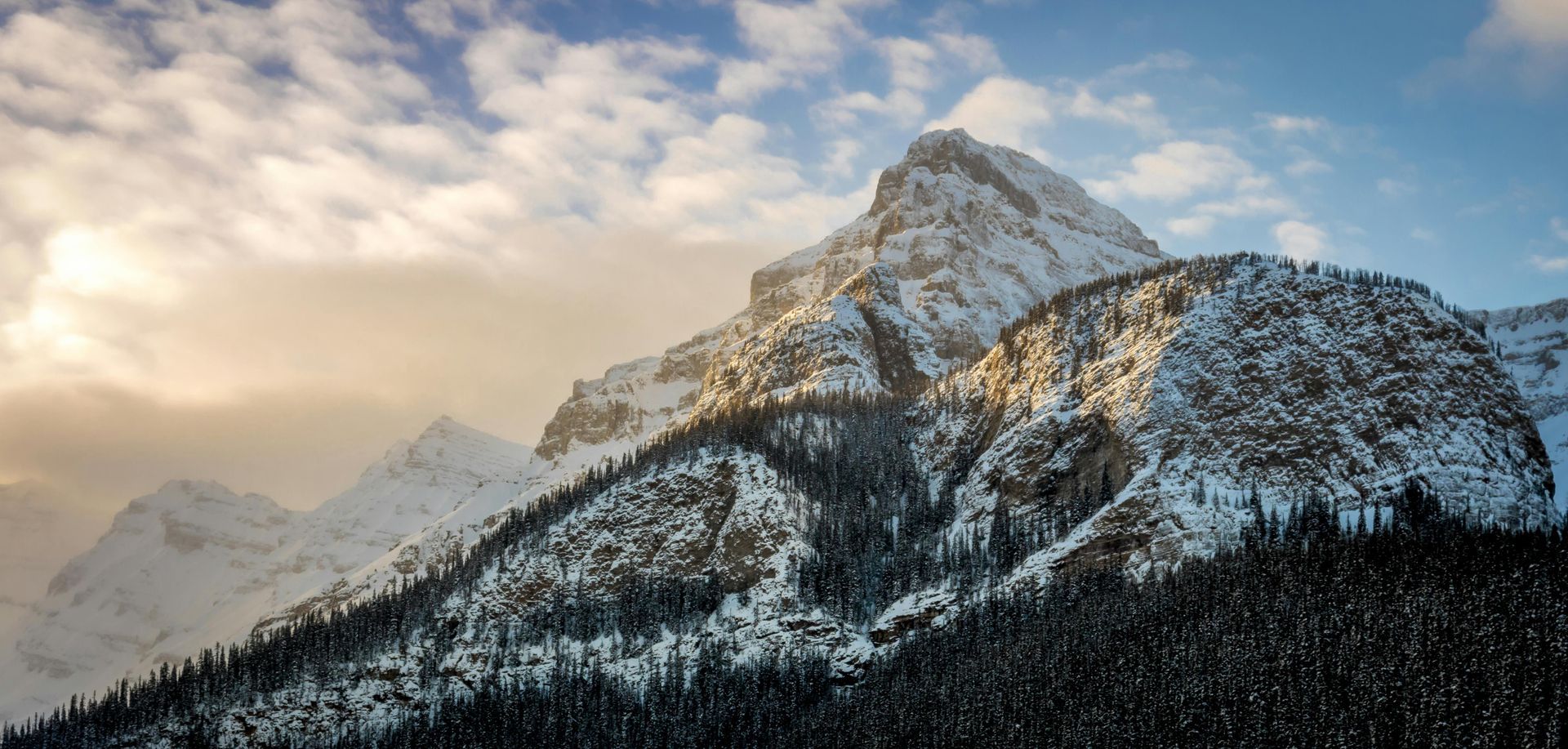 Snow-covered mountain peaks with trees below, illuminated by sunlight through cloudy sky.