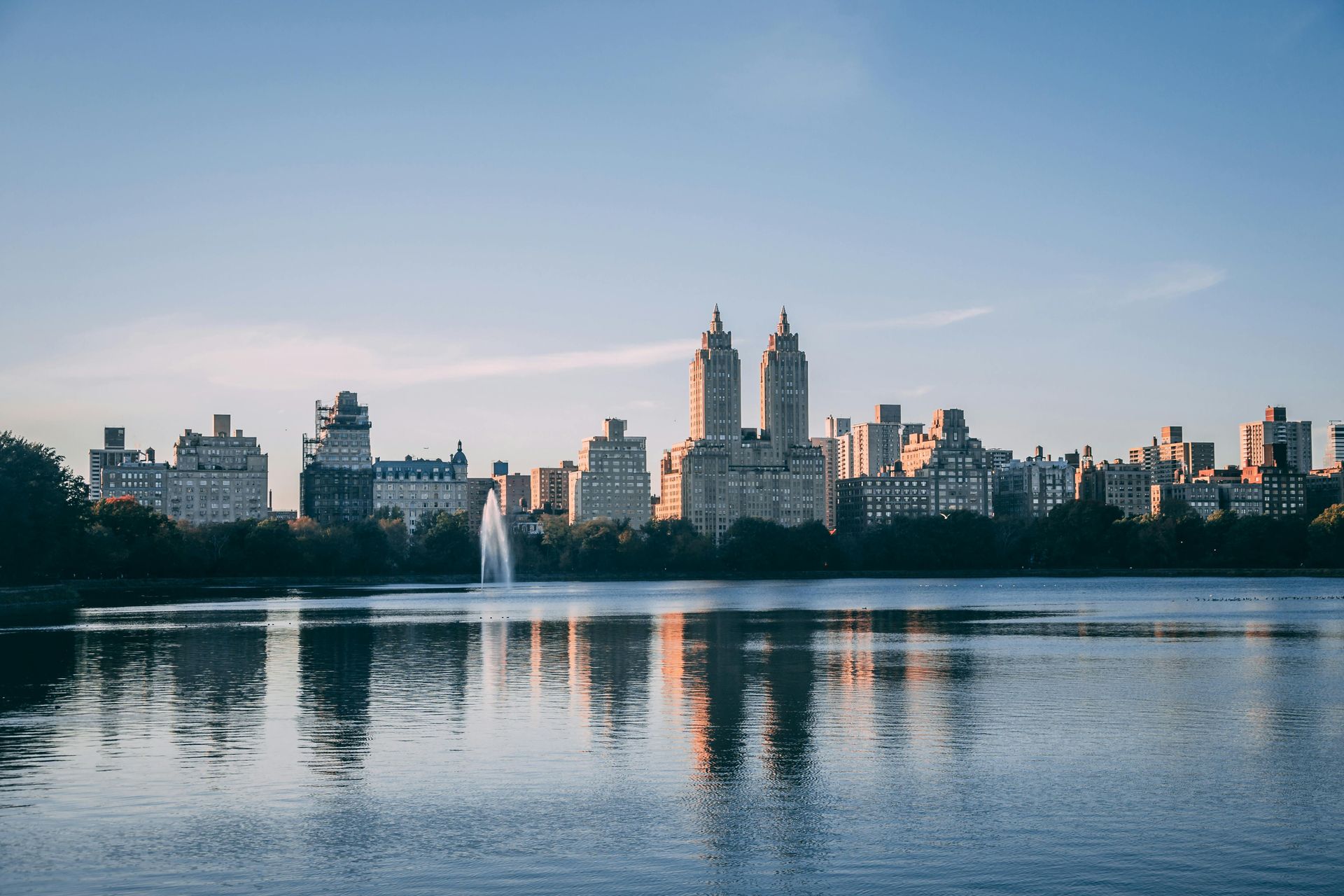 Skyline reflecting in calm water, with a fountain in the foreground and a blue sky overhead. St. Regis New York hotel central park