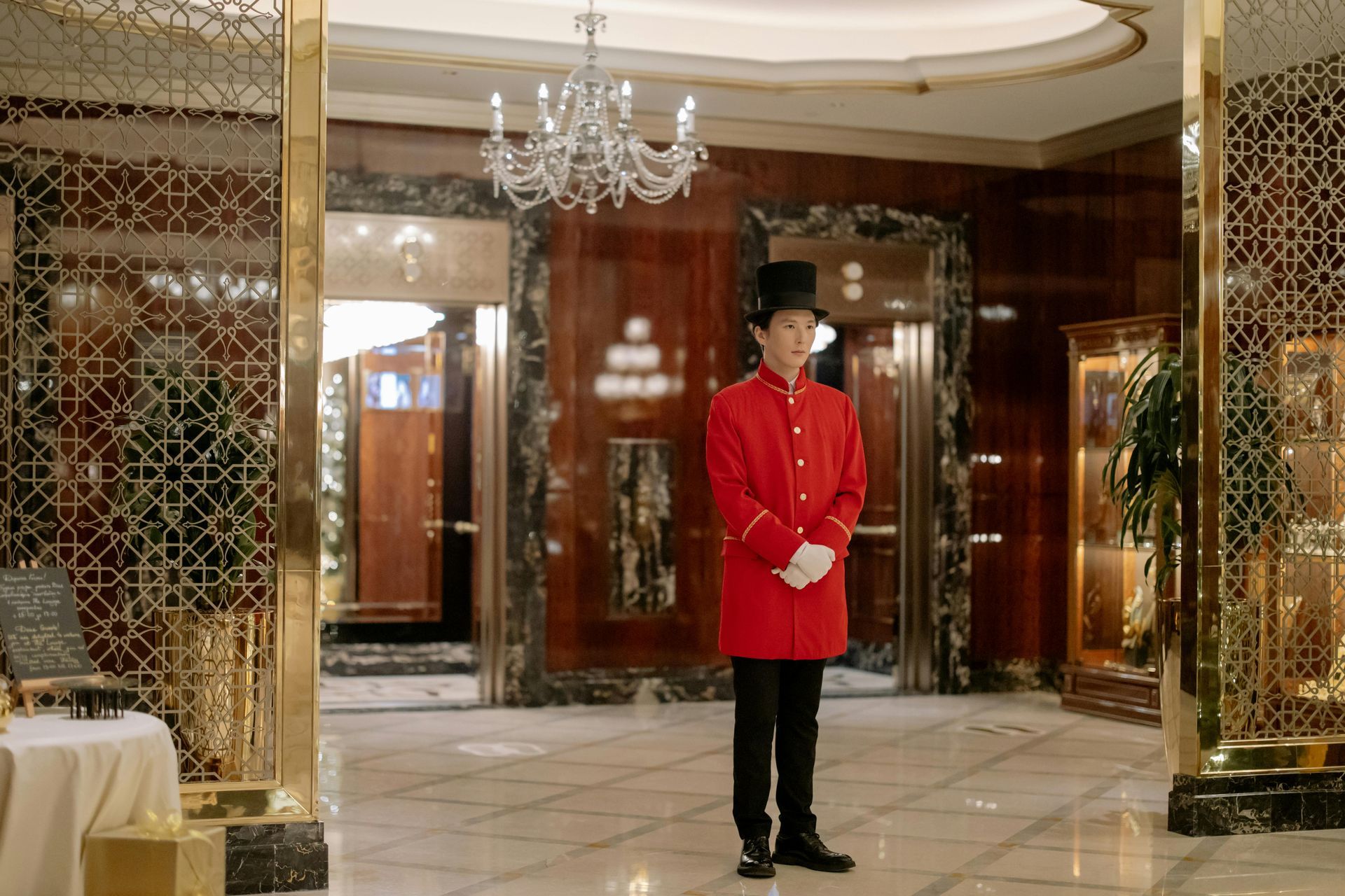 Doorman in a red coat and top hat stands in a luxurious hotel lobby with elevators and a chandelier. Tokyo hotel