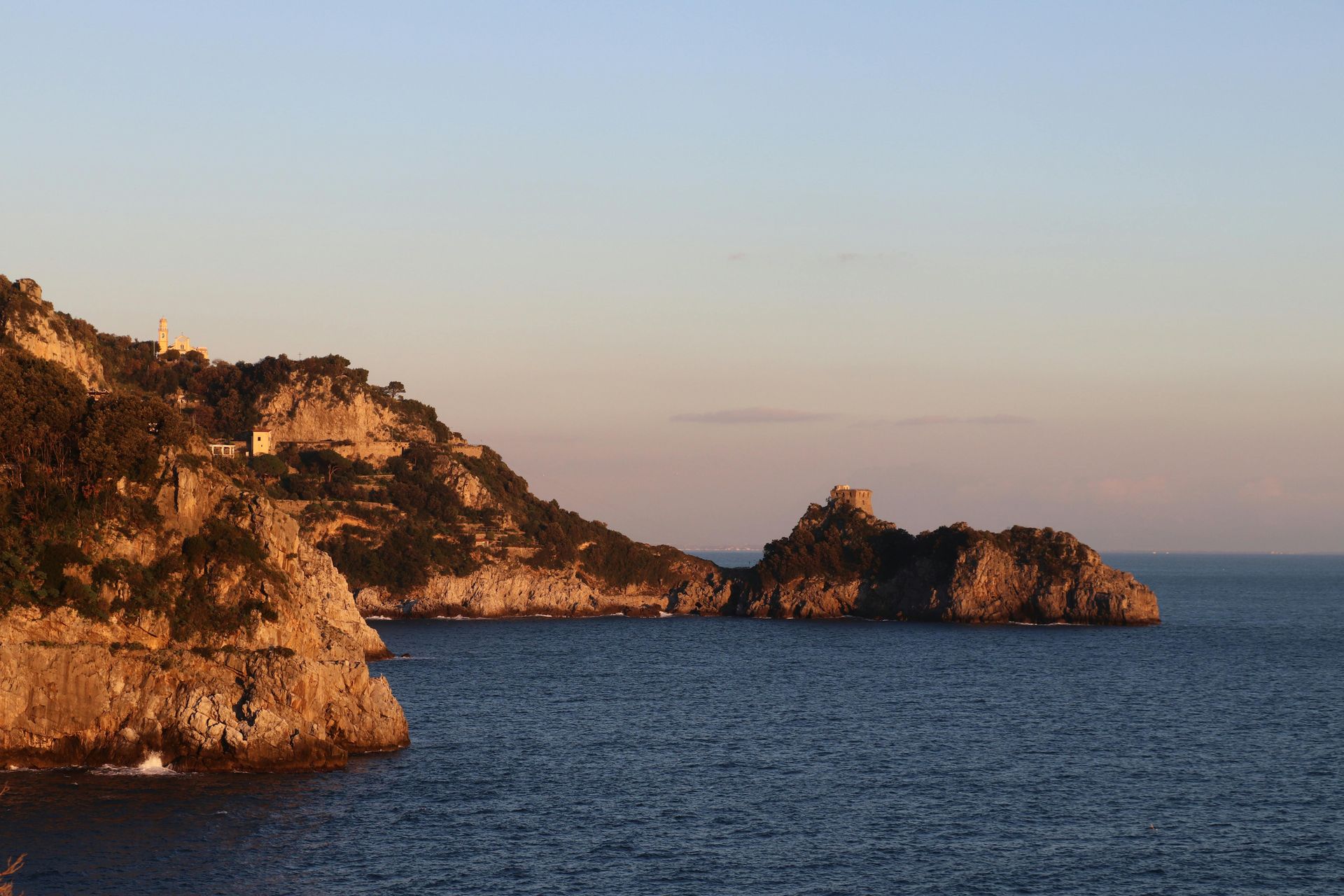 Rocky coastline at sunset, with buildings and tower, over calm blue sea. Praiano