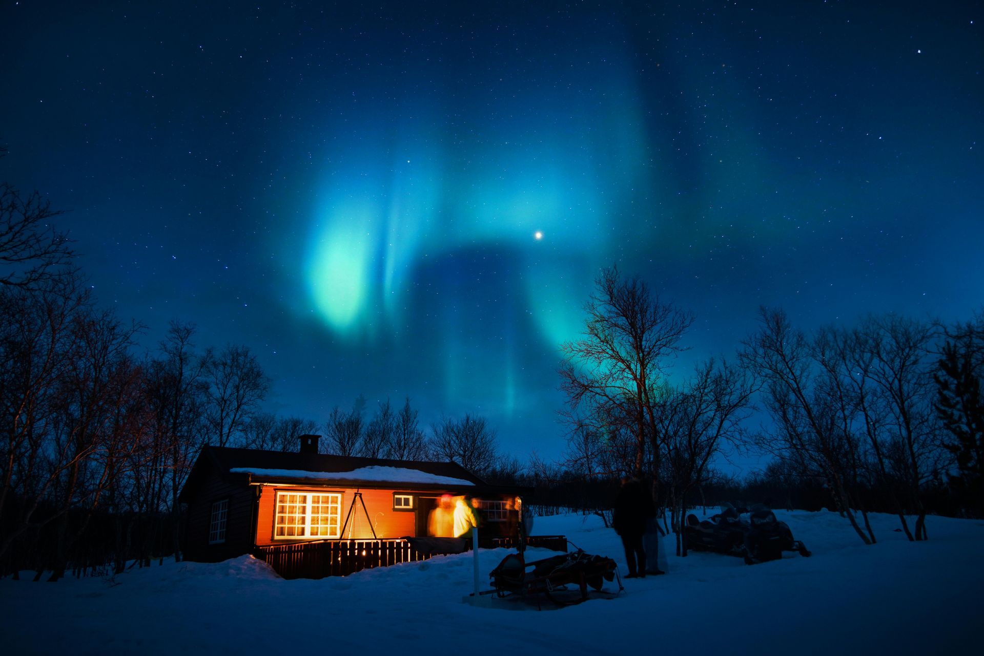 Cabin lit at night under vibrant green and blue aurora borealis.