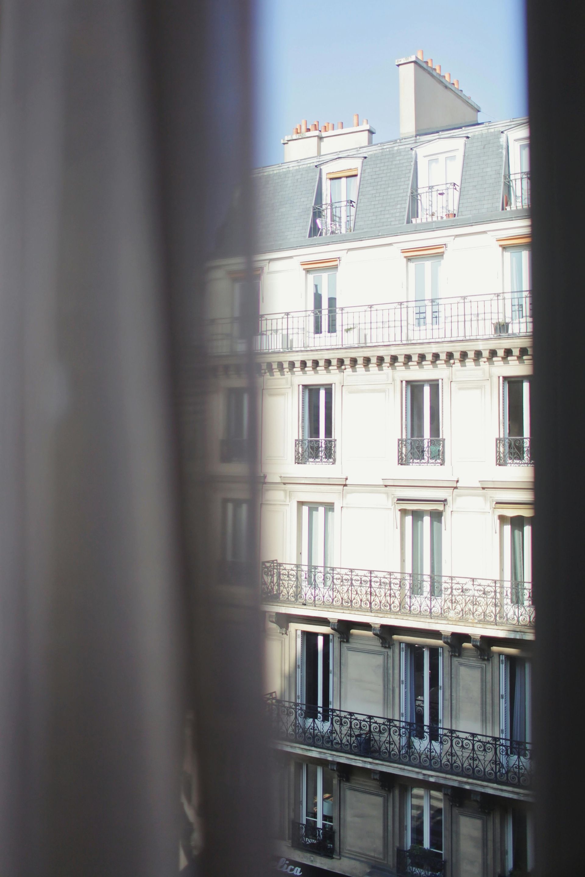 Building facade framed by a sheer curtain. Parisian architecture, cream walls, wrought-iron balconies, and gray roof.