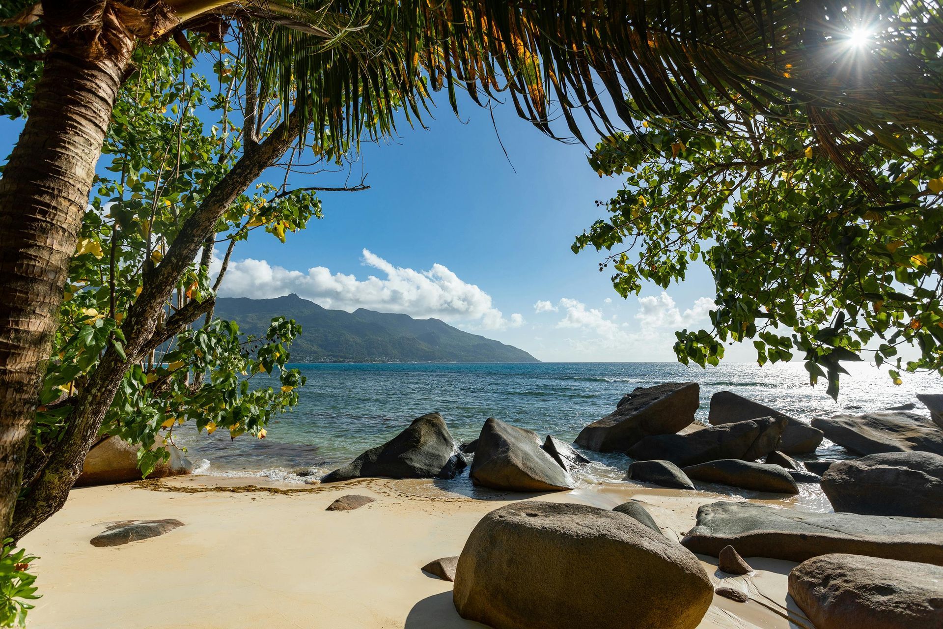 Sandy beach framed by trees, with large rocks and a view of the ocean and mountains under a bright blue sky. Seychelles