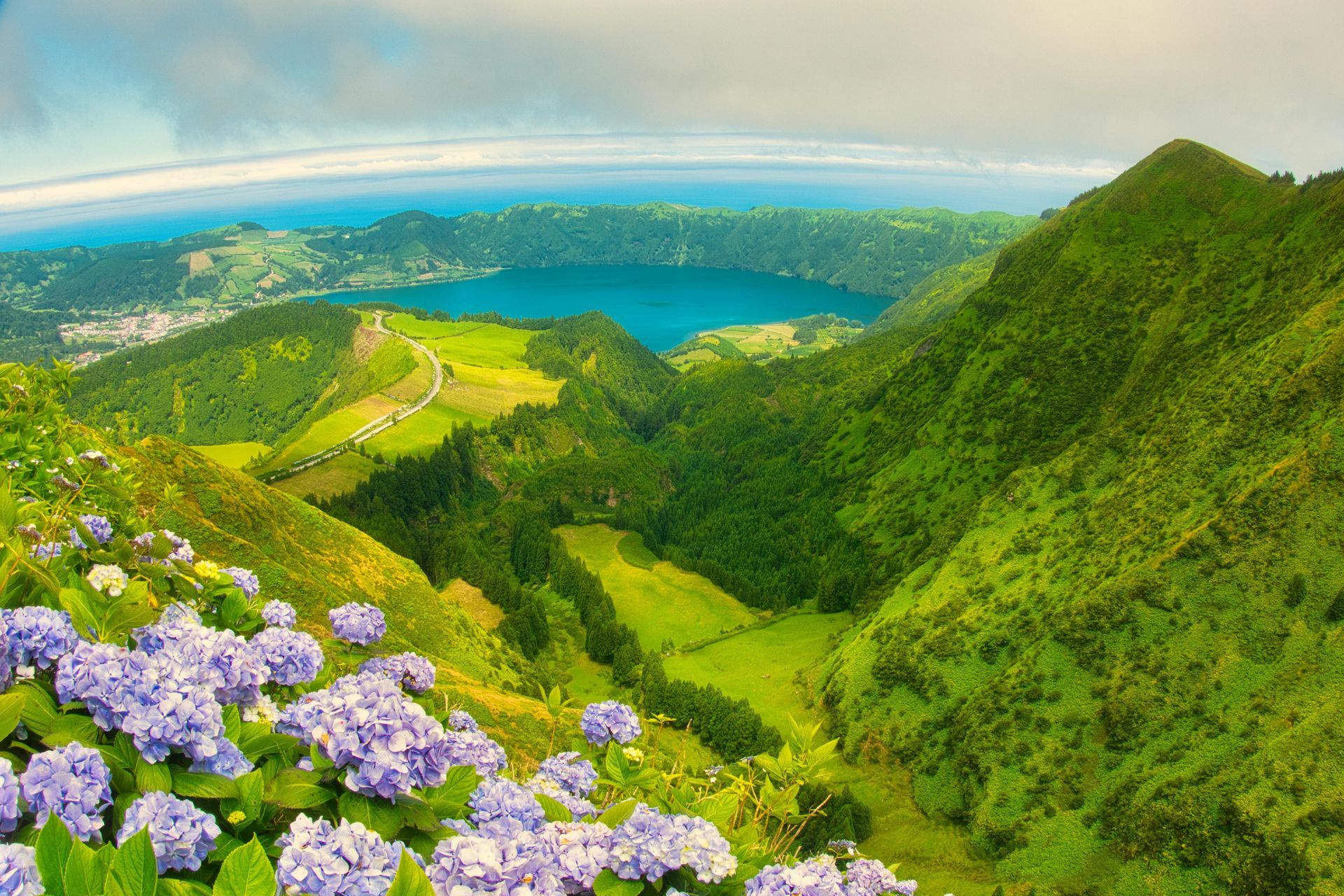 Scenic view of a lake in a volcanic crater, surrounded by lush green hills, blue hydrangeas.