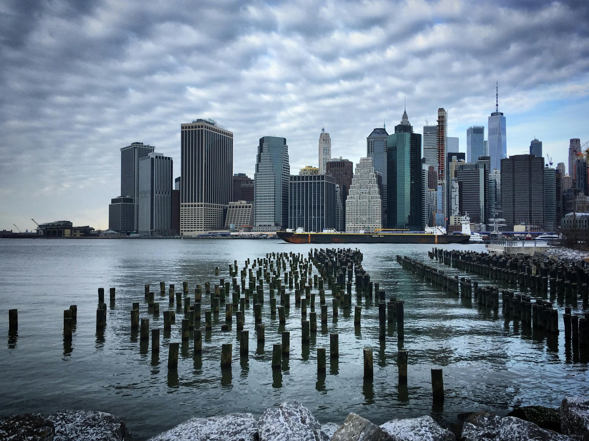 New York City skyline with cloudy sky, seen over water with old wooden posts.