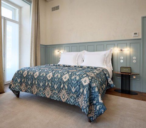 Bright bedroom with a blue patterned bedspread, white pillows, and pale blue paneled headboard by a window. Bairro Alto Hotel

