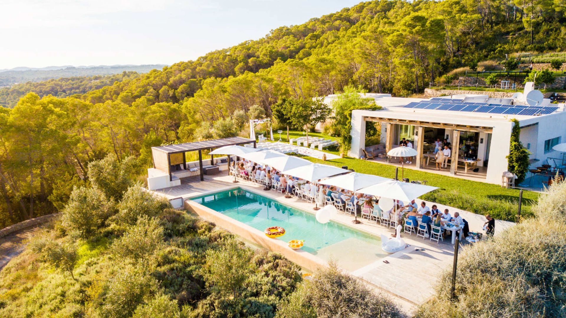 An aerial view of a villa with a swimming pool and patio, where people gather under white umbrellas on a sunny hillside.