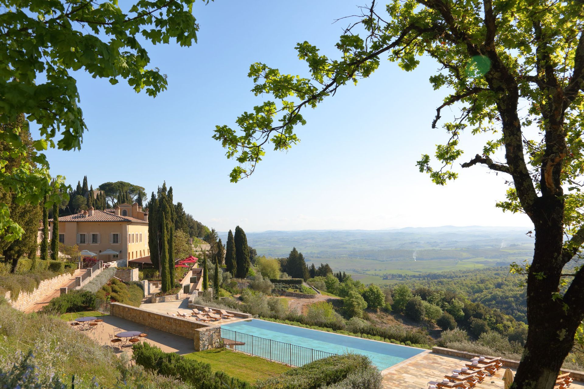 Luxury villa with pool overlooking a Tuscan valley, framed by trees on a sunny day. Rosewood Castiglion del Bosco