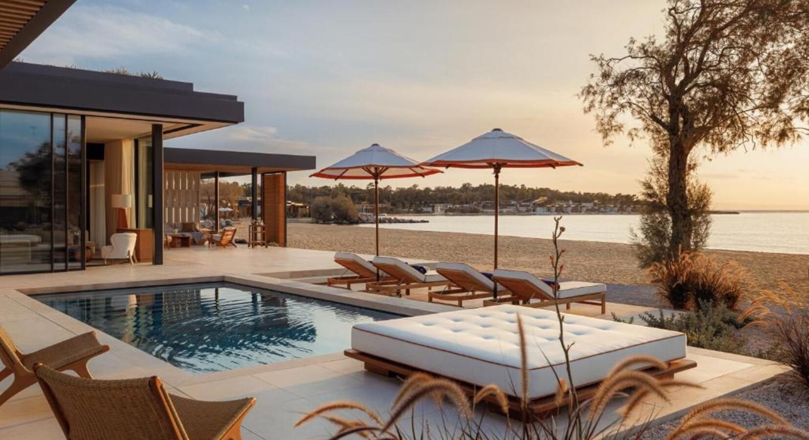 A modern house with a pool and beach lounge area, overlooking the ocean at sunset with umbrellas and lounge chairs.