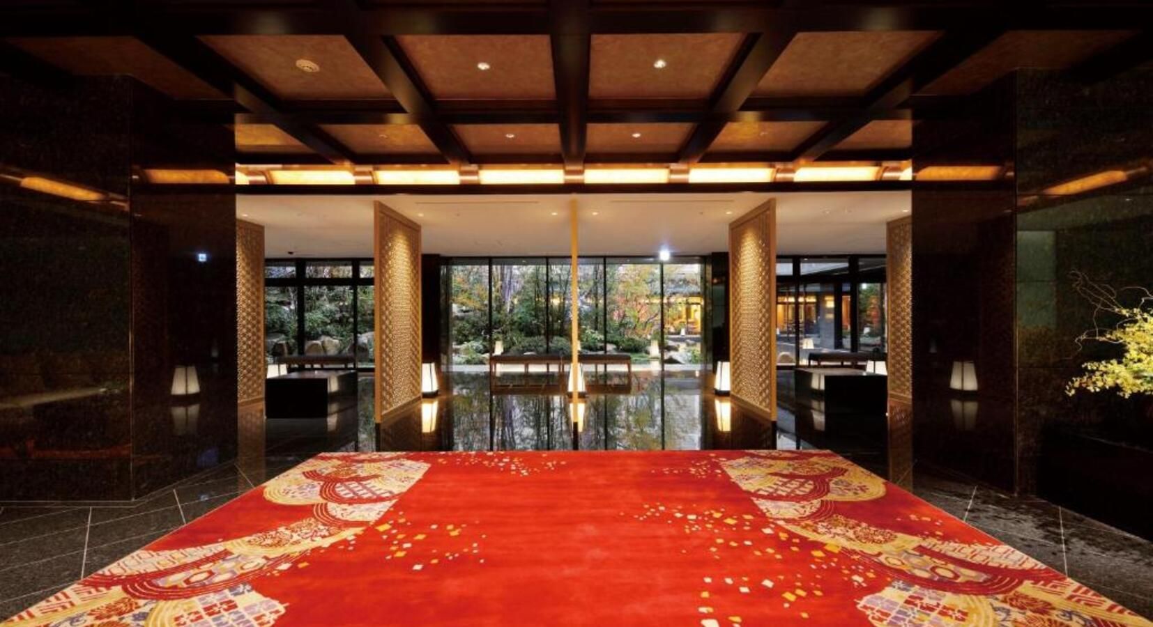 A vibrant red carpet leads through a dark, modern lobby with grid ceilings and floor-to-ceiling windows overlooking a garden. HOTEL SANRAKU KANAZAWA