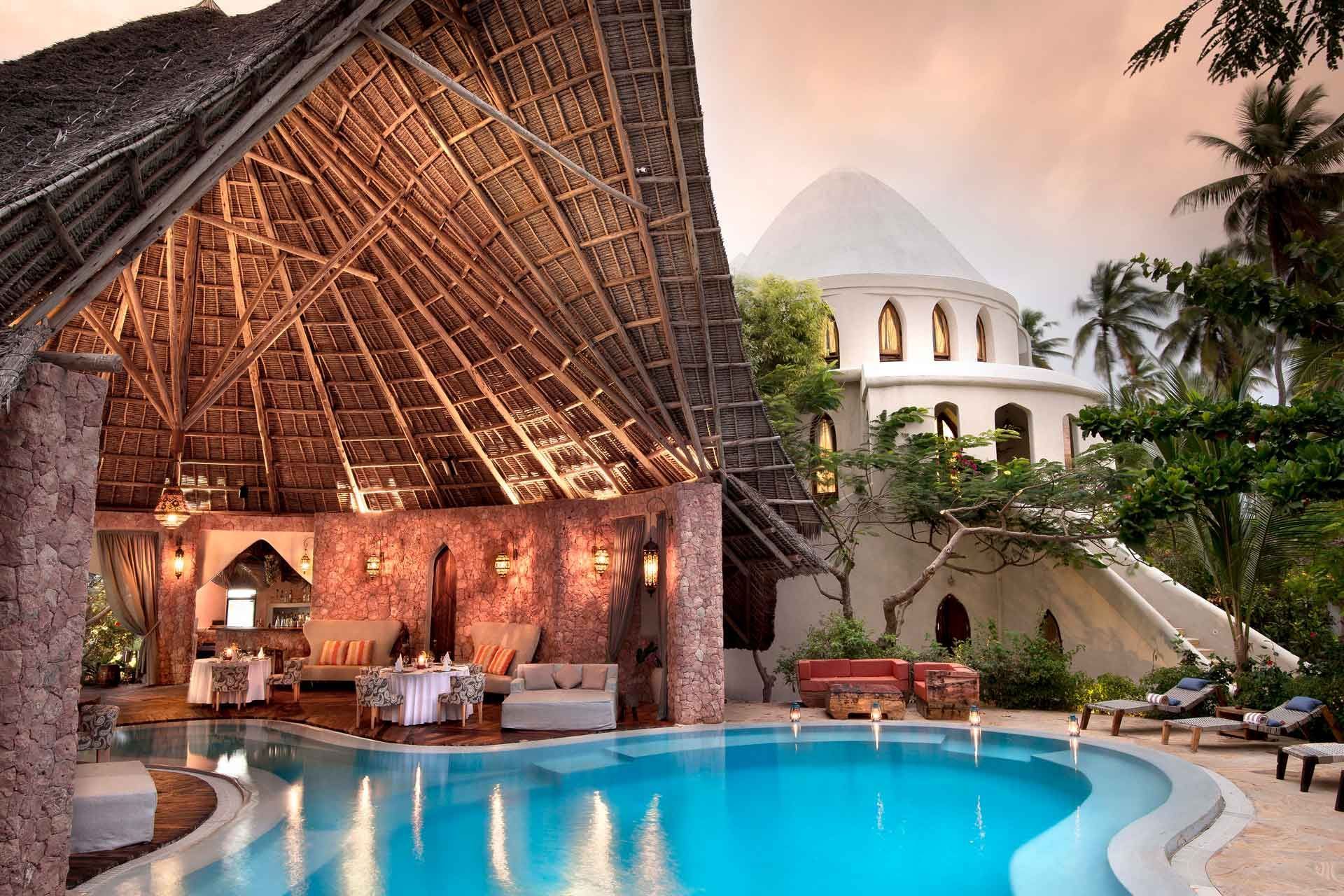 A pool area at a resort, featuring a large thatched-roof lounge, stone walls, and a white domed building nearby at sunset.