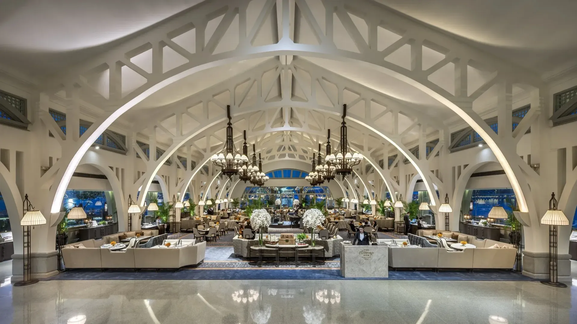 Spacious, opulent hotel lobby with high white vaulted ceilings, chandeliers, and seating areas on a polished marble floor.