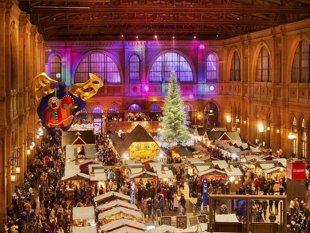 Christmas market in a grand hall with shops, people, a tree, and colorful lights.