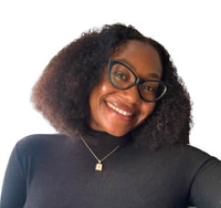 A woman wearing glasses and a necklace is smiling in front of a brick wall.
