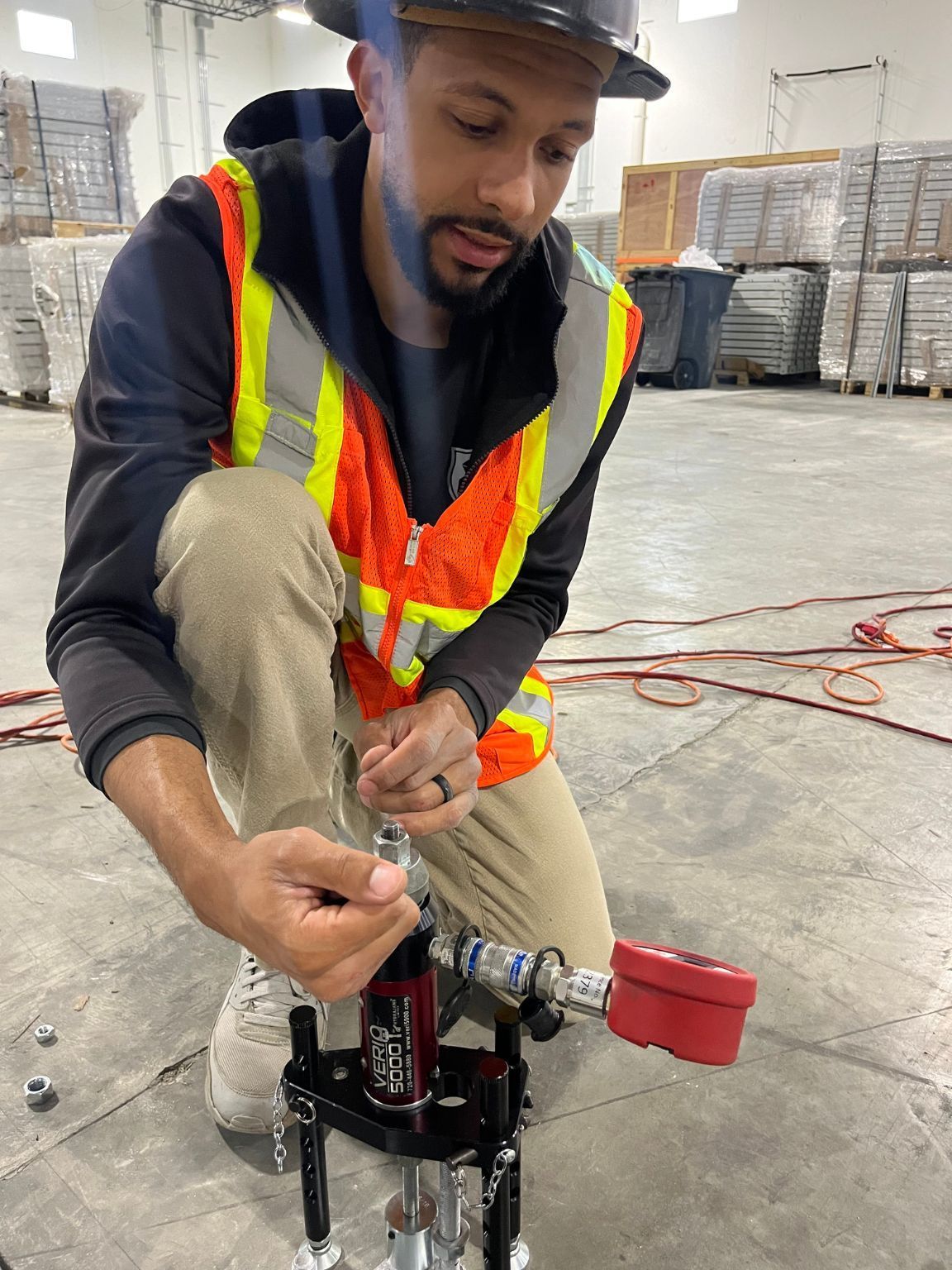 A man wearing a hard hat and safety vest is working on a machine in a warehouse.