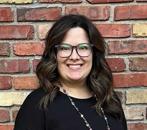 A woman wearing glasses and a necklace is smiling in front of a brick wall.
