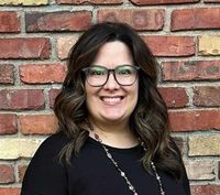 A woman wearing glasses and a necklace is smiling in front of a brick wall.