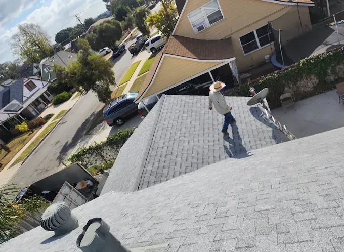 Person in hat walks on a gray shingle roof, suburban street in background.