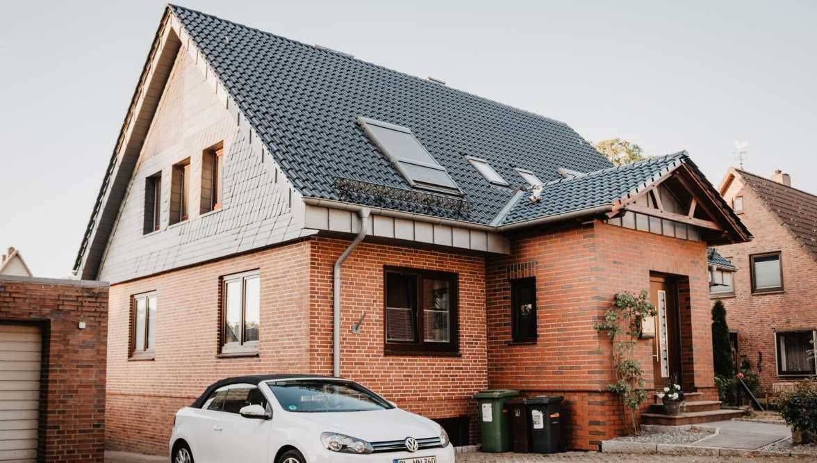 Brick house with gray roof, white car parked in front, green trash cans on the side.
