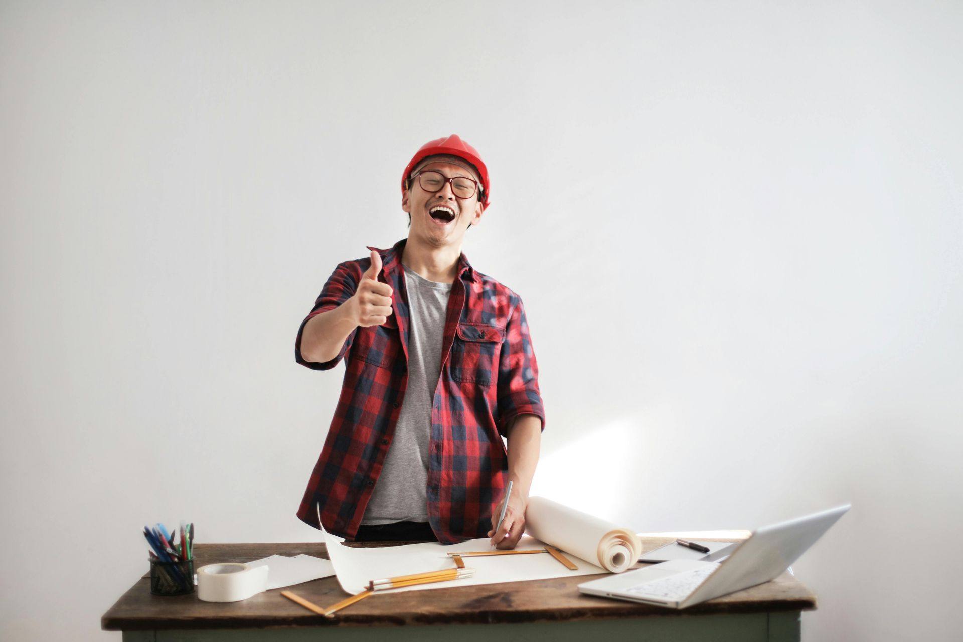 Man in red hat and flannel shirt smiles and gives a thumbs-up at a desk with blueprints and laptop.