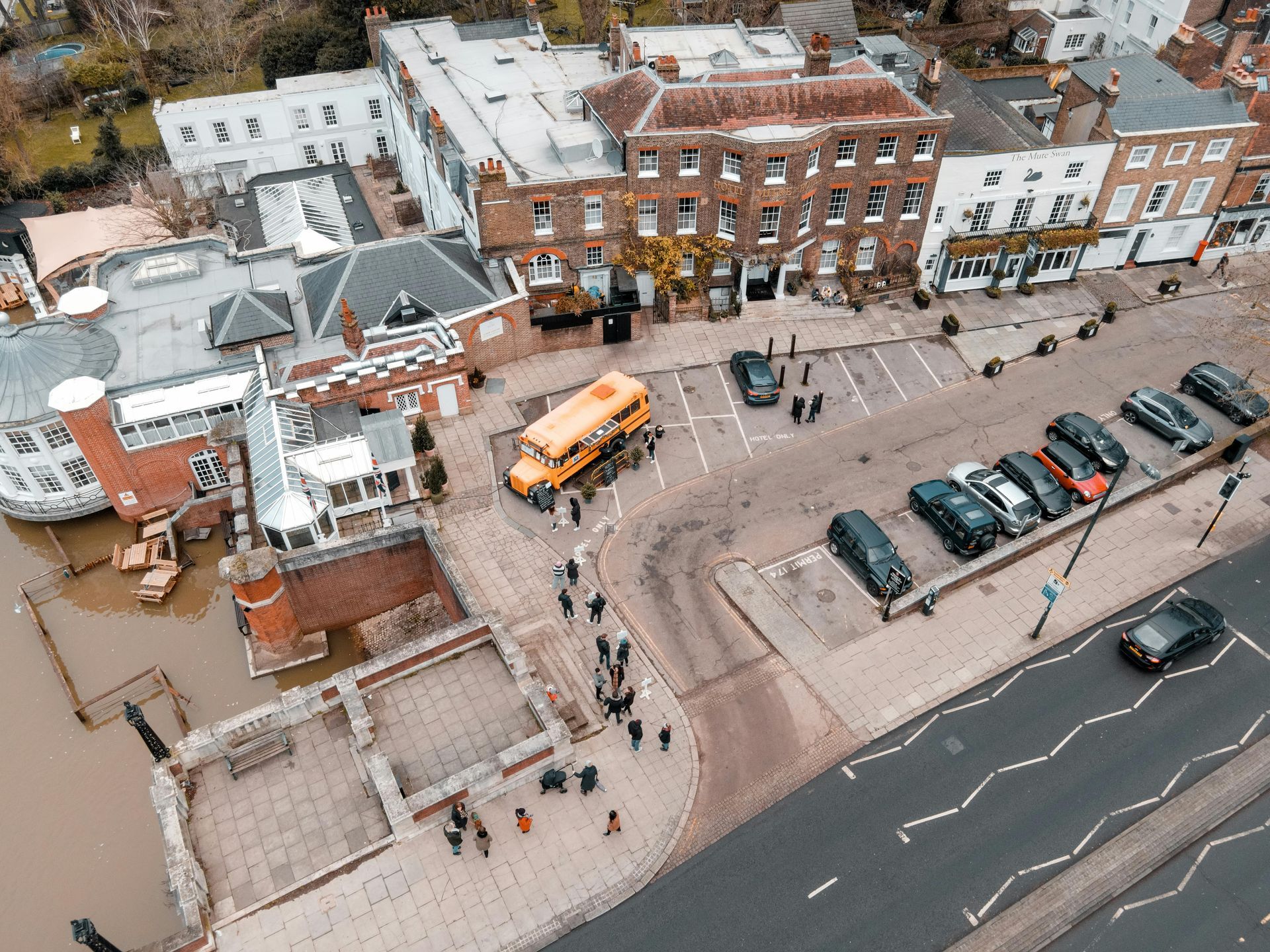 Aerial view of a brick building, a yellow school bus, and parked cars on a street near the water.