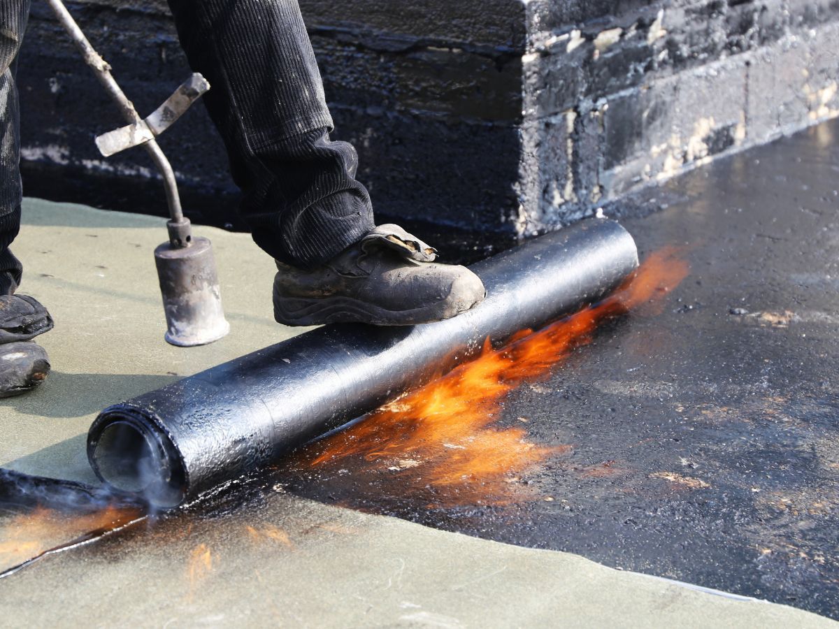 Person using a torch to apply a rolled roofing membrane. Flames and dark material are visible on the roof.