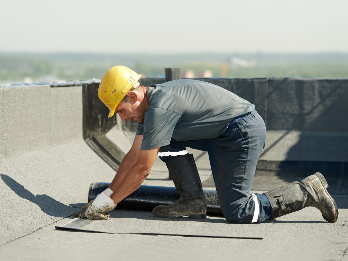 Roofer kneels, applying sealant on a flat rooftop, wearing a yellow hard hat and work gloves.