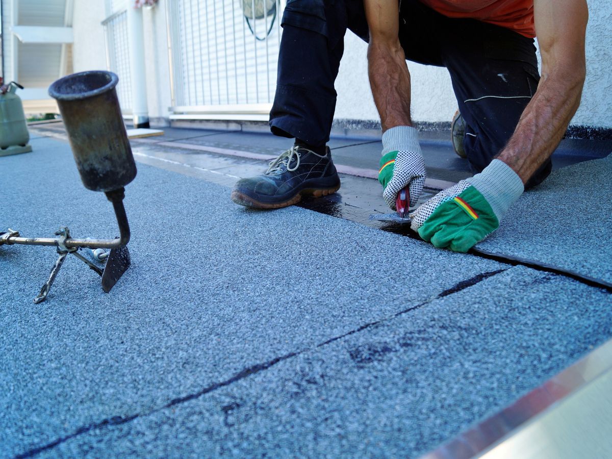 Roofer using a torch and knife to repair a flat roof, wearing gloves and work boots.