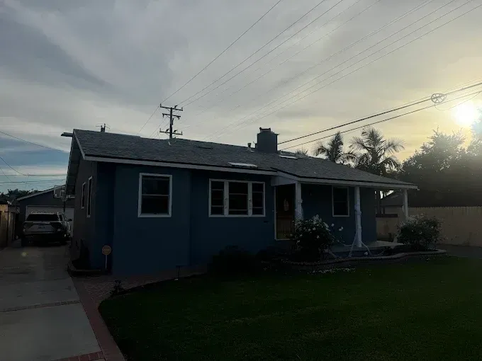 Blue house with white trim, green lawn, driveway, and palm trees against a cloudy sky.