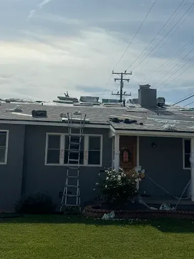 House with damaged roof and ladder, debris scattered. Power lines and cloudy sky in background.