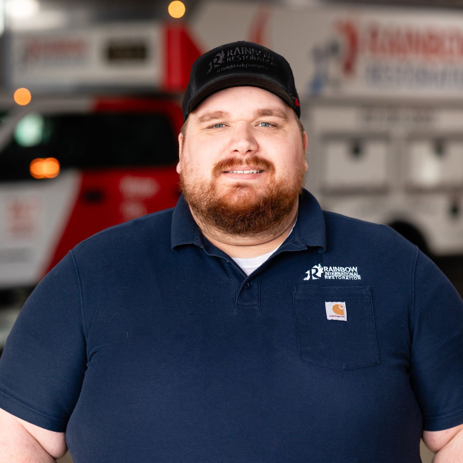 A smiling man with a beard is wearing a hat and a blue Rainbow Restoration shirt.