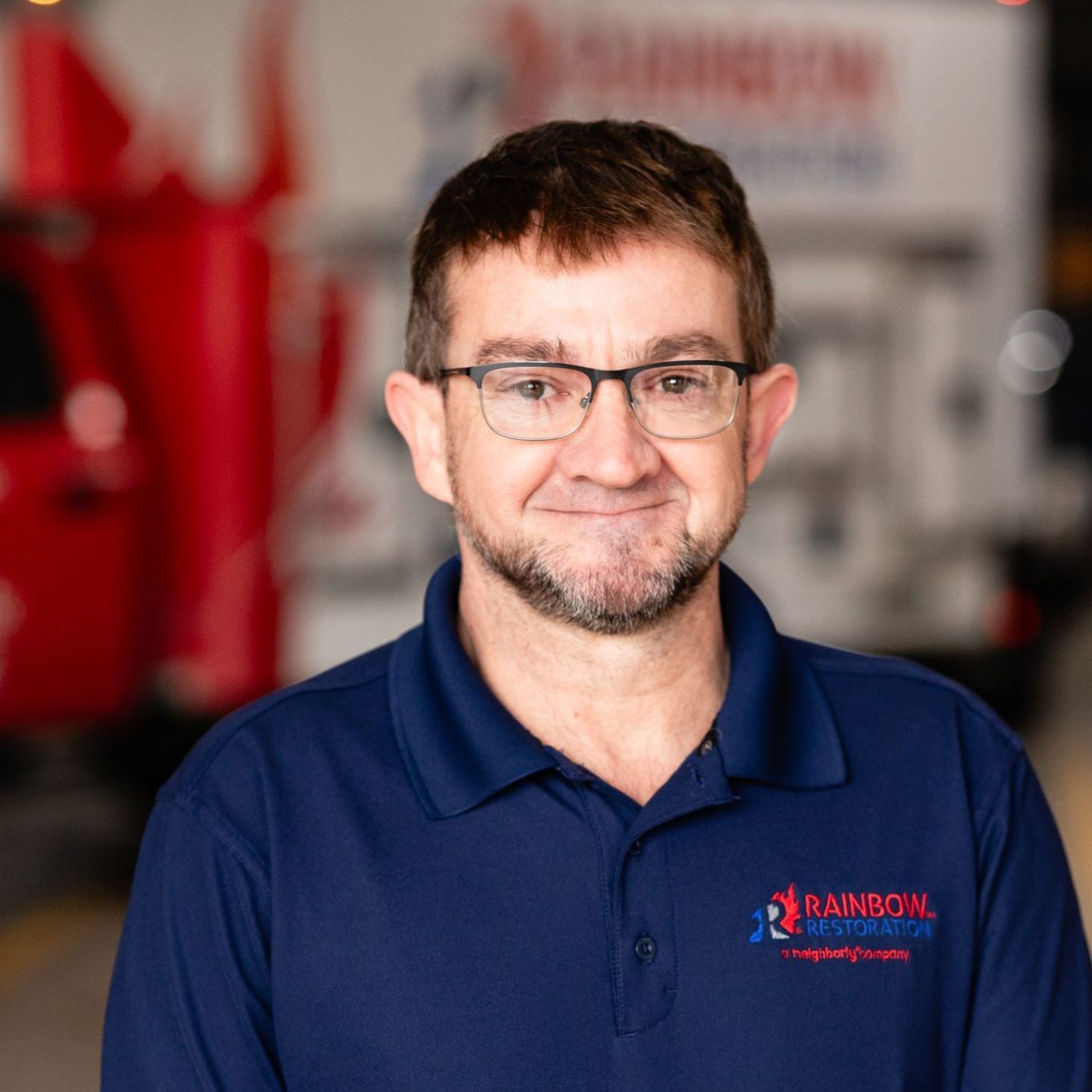 A smiling man wearing glasses and a blue Rainbow Restoration shirt is standing in front of a red truck.