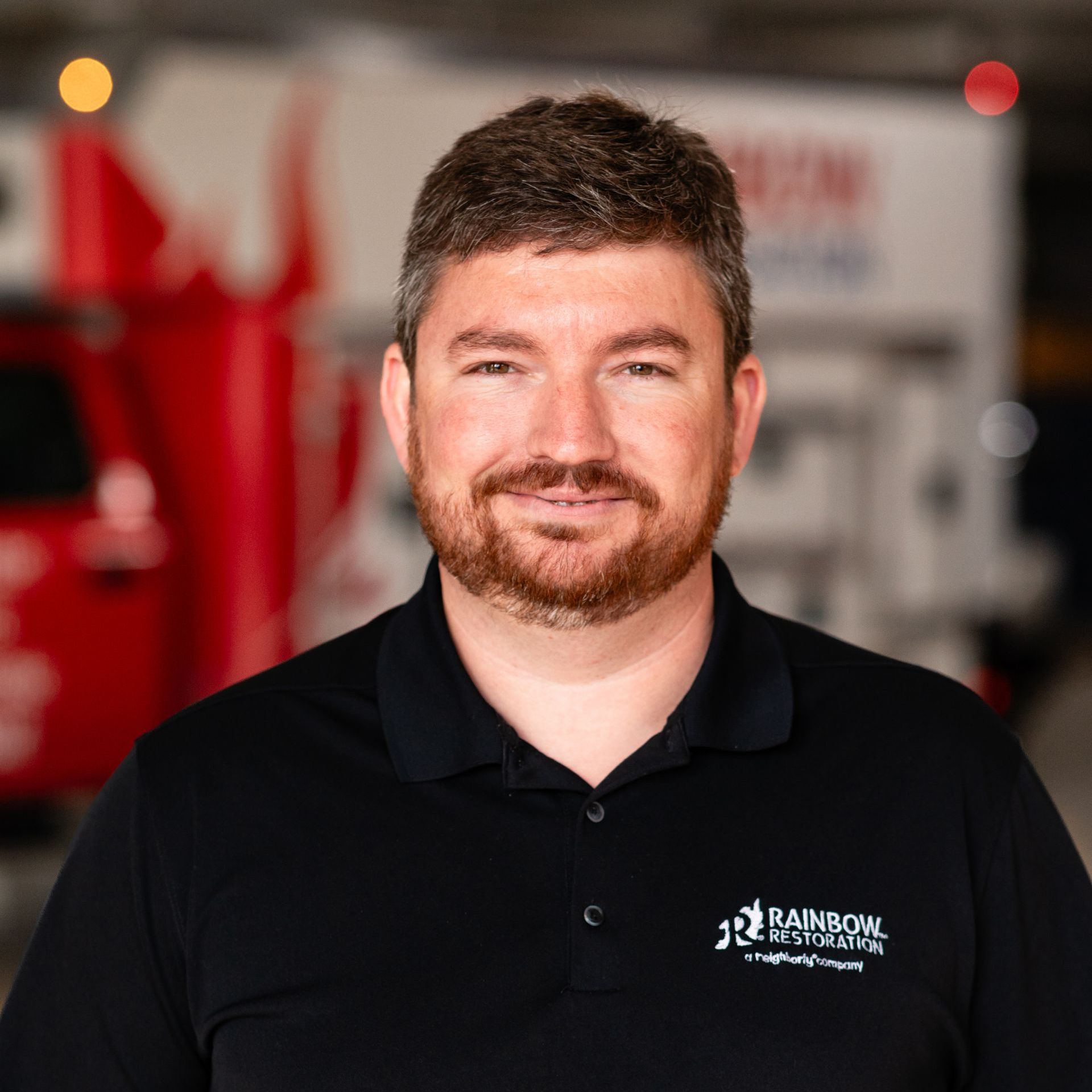 A man with a beard is wearing a black shirt and smiling in front of a red truck.