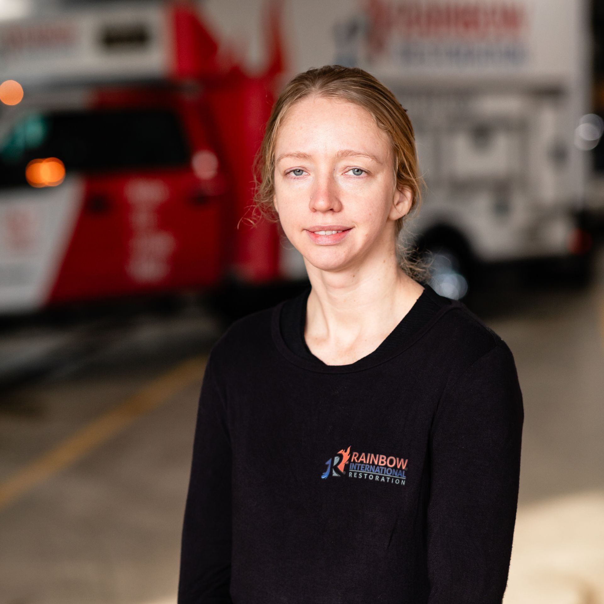 A woman wearing a black Rainbow Restoration shirt is standing in front of a truck smiling.