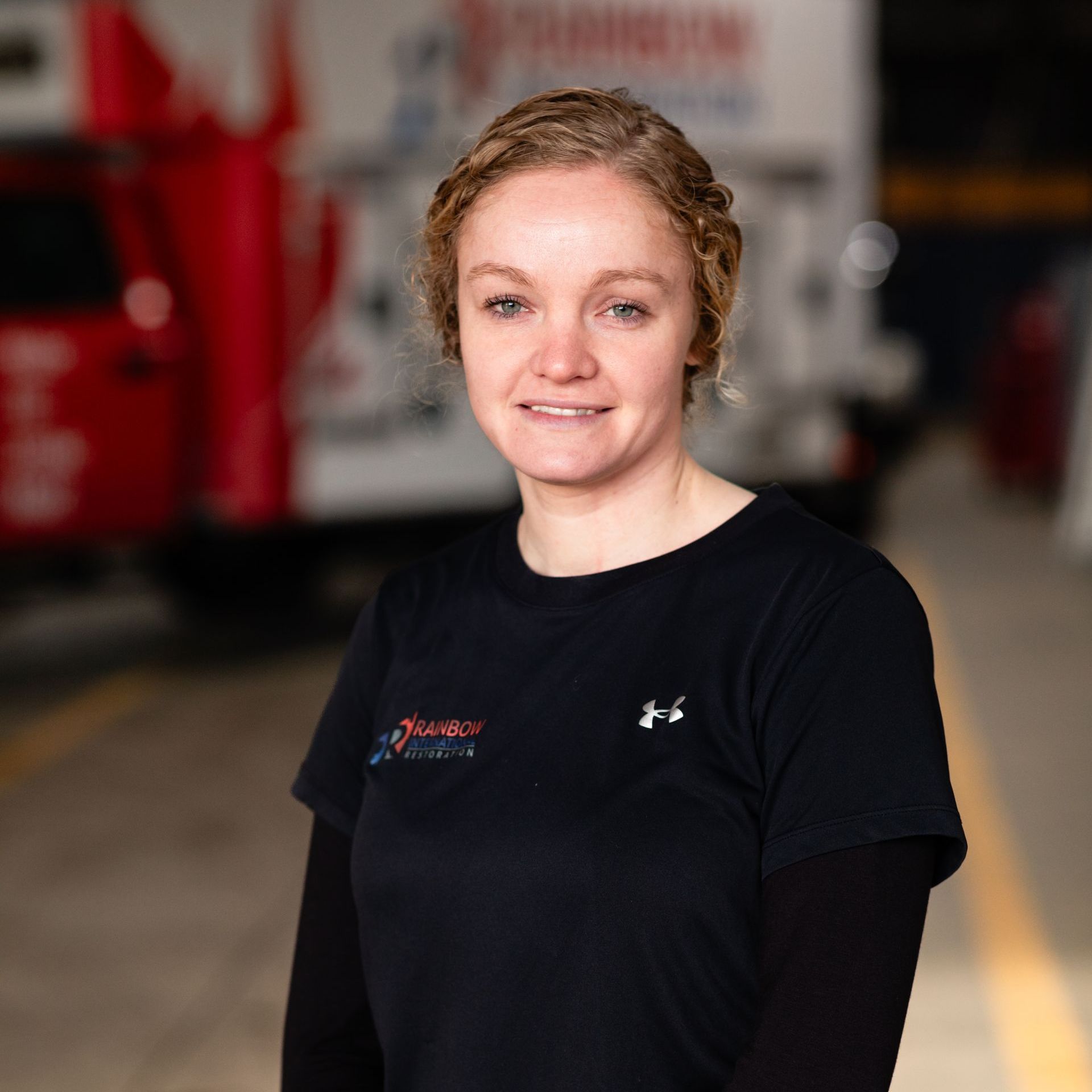 A woman in a black Rianbow Restoration shirt is standing in front of a truck smiling.