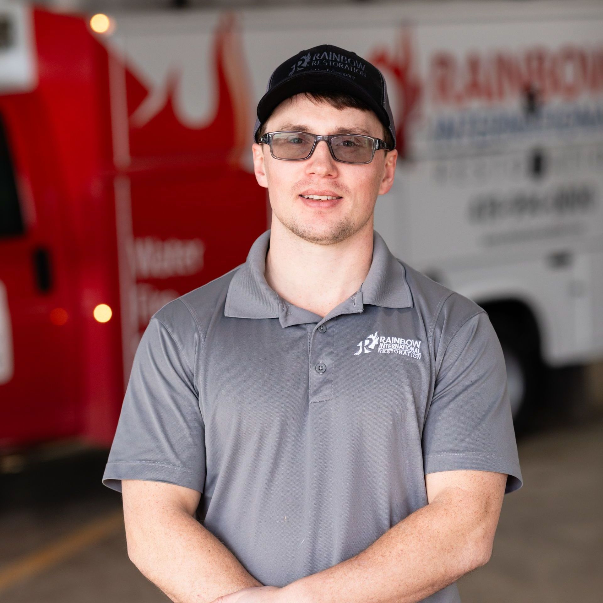 A man wearing a hated gray Rainbow Restoration shirt is standing in front of a red truck smiling.