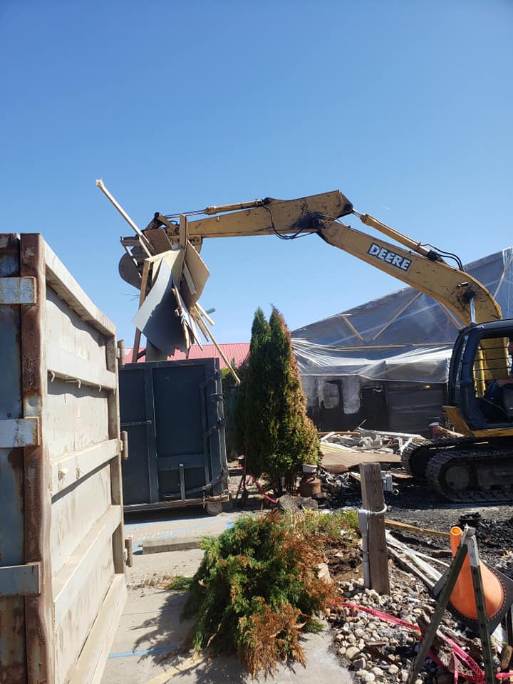 A large yellow excavator with the word Deere on it is emptying a load of rubble into a dumpster.