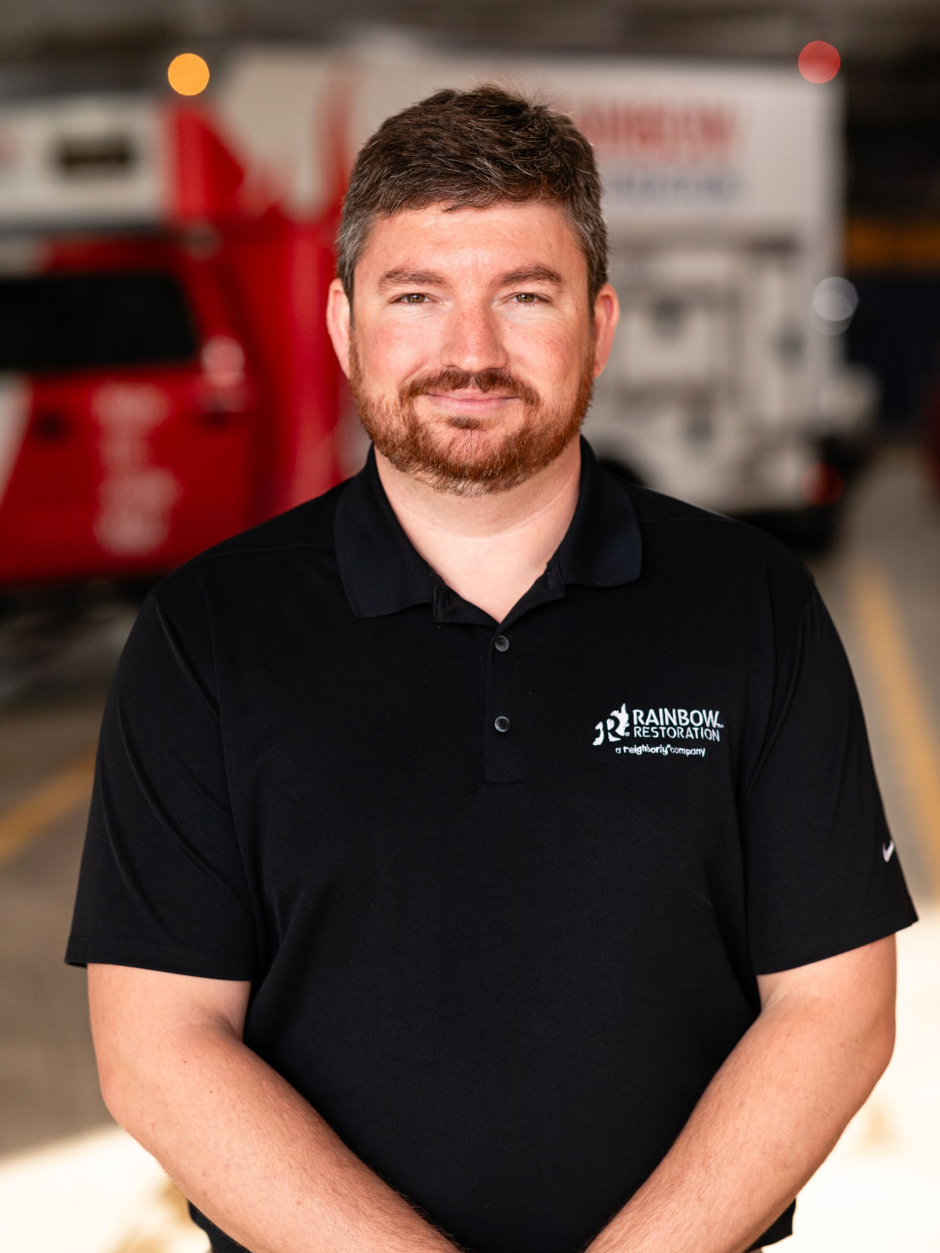 A smiling man with a beard is standing in front of a red truck in a parking garage.